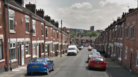 A street with terraced houses on either side of the road, with cars parked on both sides and more terraced properties in the distance