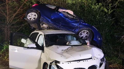 The scene of a car crash, with a blue car piled on top of a white car, both badly damaged