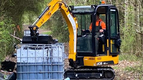 A JCB digger putting contaminated soil into a plastic cube in the woods