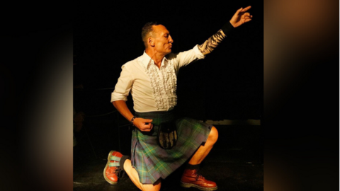 Piers Burnell dressed in a kilt, kneeling down and poses for an air guitar performance. He is in profile and his eyes are closed.