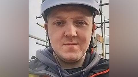 A man looking into the camera while wearing a light blue or grey hard hat, seemingly on a building site. 