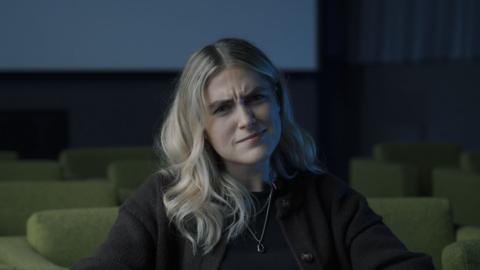 A woman with blonde hair and a quizzical look sits alone in a lecture hall with green chairs.