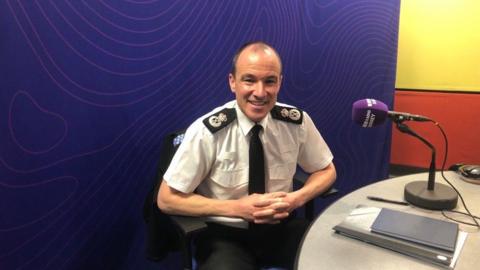 Chief Constable Tim De Meyer sits in the BBC Radio Surrey studios. He is wearing a white shirt and black tie. There is a purple board behind him and a purple microphone, with the words 'BBC Radio Surrey' on it. A notebook and folder are on the table in next to him.