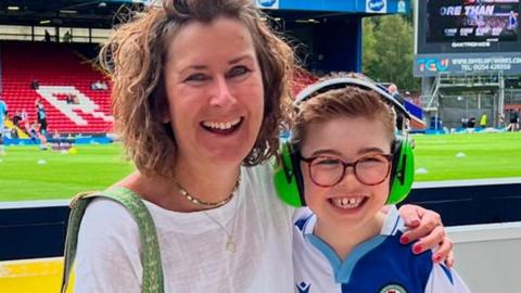 Debbie Allingham beside the pitch at Blackburn Rovers with her arm round her son, who is wearing a Rovers shirt, glasses and headphones. Both are smiling