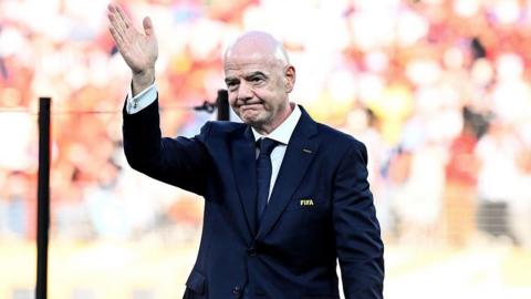 Gianni Infantino waves while wearing a dark blue suit with a Fifa pin