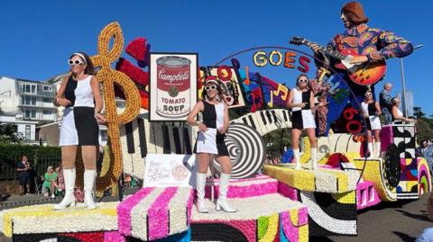 A giant figure of a guitarist in a blue shirt with orange paisley pattern is at the end of a float with a black and white keyboard. There is a multi-coloured sign reading Pop Goes the 60s and four women are standing on the float. They are wearing sleeveless mini dresses with black and white opposite rectangles. 