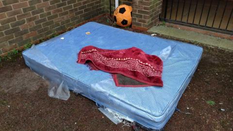 A light blue and white striped mattress with some ripped plastic wrapping around it is dumped on the concrete ground outdoors. There is a red blanket with a white geometric print around the edging which has been left on top of the mattress and an orange and black large inflatable football in the background.