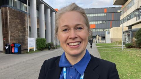 Mae Dr Munro yn sefyll y tu allan i Brifysgol Aberystwyth, gydag adeiladau a glaswellt y tu ôl iddi. Mae hi'n gwisgo blows las a siaced ddu ac yn gwenu.