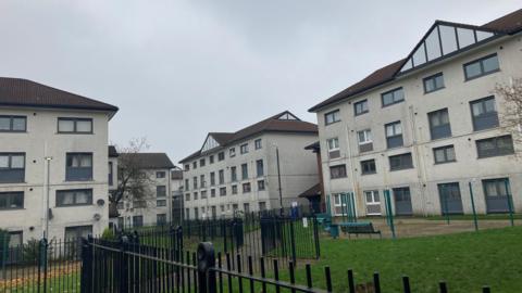 Blocks of flats in the background, with black metal railings in the foreground and small green fields. 