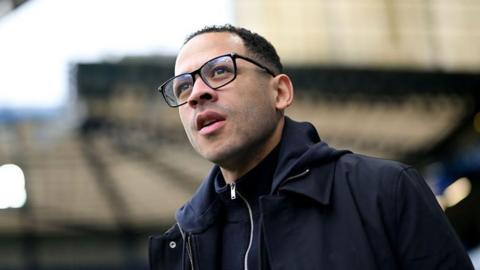 Close-up of Liam Rosenior. He has short black hair and is wearing black-rimmed glasses and a navy coat.