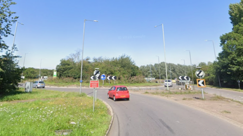 Cars are approaching and circling a roundabout, which has grass and bushes on it. It is a sunny day with blue sky above.
