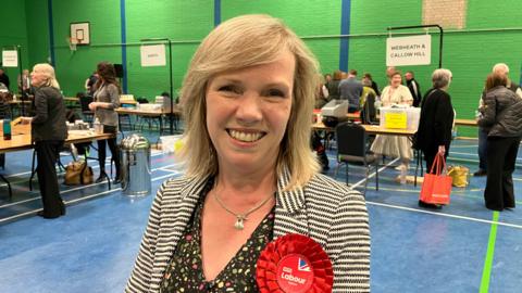 Labour Councillor Sharon Harvey, the leader, is stood at the election count wearing a red Labour rosette. Vote counting staff are standing at a number of tables in a sports hall that has a blue floor and green walls.