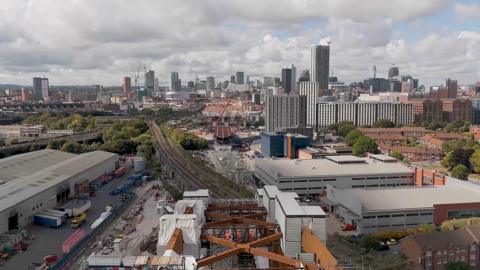 An aerial view showing the construction of a section of railway viaduct, with struts holding it up and workers in orange fluorescent uniforms. Tall city buildings can be seen in the distance on a cloudy day. 