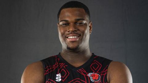Anthony 'TJ' Hibbert in a black basketball vest with a red pattern and Essex Rebels logo. He smiles at the camera in front of a grey background. 