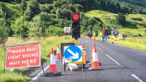 A traffic light, on red, traffic cones and signs sit on a two-lane country road on a sunny day. Maintenance workers wearing high-visibility gear, as well as construction vehicles, can be seen in the distance. The road side is lined by green grass, with trees and bushes on the hills in the background. 
