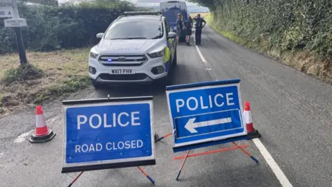 a police vehicle is parked on a country road behind two police cordons.