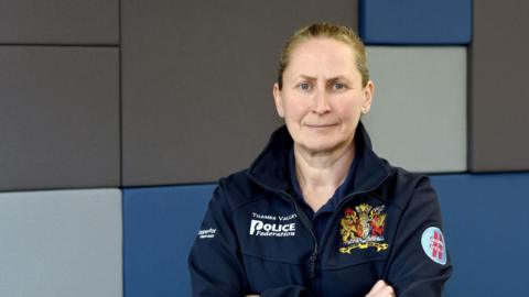 Aileen O'Connor stood with her arms crossed. She has brown hair that is tied back, and is wearing a coat with the name of the police federation and its logo embroidered in. The wall behind her is blue and grey.