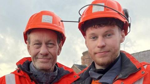 Two men dressed in orange hi-vis clothing with a logo that reads Hydro and wearing orange hard hats stand in a walled outsdie space in front of some oak benches. One of them is a man in his 50s and the other, a younger man of 21 with a short ginger beard.