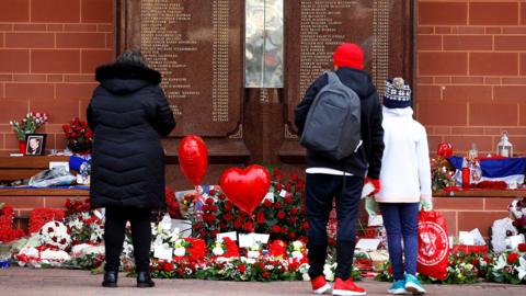 People stand in front of the Hillsborough Memorial outside Anfield Stadium in Liverpool. There are flowers and heart balloons in front of a plaque with the names of the victims of the disaster.