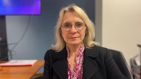 Elaine Millar - A woman with shoulder length blonde hair wearing glasses, a black blazer jacket with a pink floral shirt underneath. She is sat at a desk.