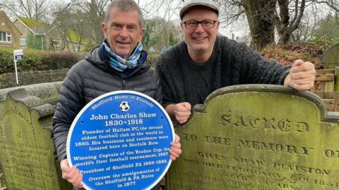 A man wearing a black jacket and a scarf holds a blue plaque. He is stood next to an old headstone. Another man is stood behind the headstone and wearing a flat cap, glasses and a dark grey jumper.