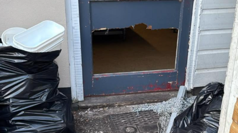 The image shows the entrance to a business or commercial premises where the lower panel of a blue-grey door has been smashed or kicked through, leaving a large rectangular hole. Broken glass is scattered on the ground outside the doorway, and black bin bags can be seen around the entrance.