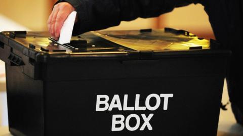 Image shows a black ballot box with a hand putting a voting paper in