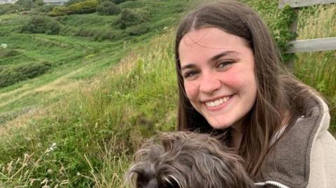 Maddy Lawrence is looking at the camera smiling with a dog on her lap. There is a sweeping green hillside in the background.