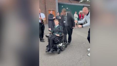 A teenager in a wheelchair, in a playground. Behind him are numerous teachers and friends.