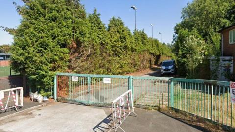 Locked green entrance gates to the site, with a row of conifers along the path and a van parked on the drive 