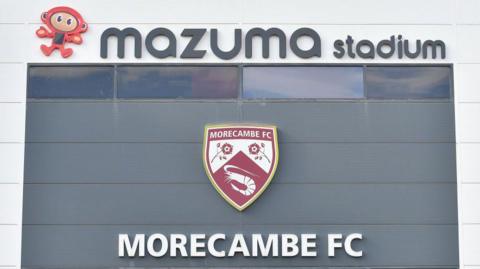 A view of the outside of Morecambe's Mazuma Stadium with the badge and 'MORECAMBE FC' emblazoned on it