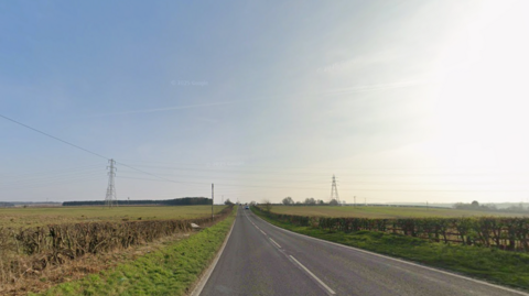 A country road. Green fields are on each side of the road. Pylons can be seen in the distance. The sky is blue.
