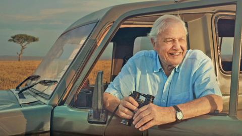 Sir David Attenborough leans outside of a safari van holding a pair of binoculars.
