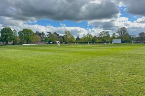 A wide green cricket field under a partly cloudy sky, with a pitch and trees lining the background.