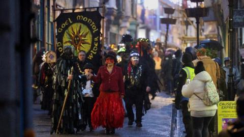 A group of people walking down the street holding a sign which reads Goel Montol, one of the people is wearing a red mask, hat and red jacket and dress, others around them are wearing costumes and masks.