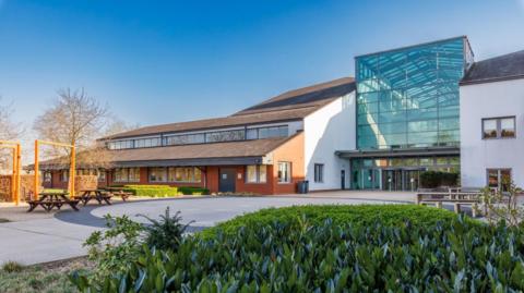 A large office building with a middle section which has a tall glass frontage. The section to the left has a sloping roof on two levels, and to the right there is a standard two-storey building. There are benches on the pavement area in front with plants in a bed.