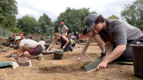 A group of people, some young students, others retired volunteers work on their hands and knees removing soil from a graveyard at the dig site.