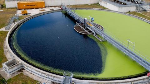 An aerial photos of a sewage treatment works, showing a round filtration pond half covered with green algae on the right. Surrounding the pond is a grass area and some smaller tanks to the right