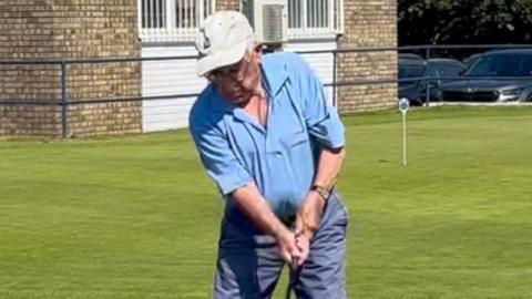 An elderly man getting ready to swing a golf club. He wears a cap, blue polo top and navy blue shorts.