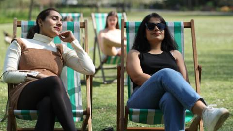 Two young women relax in park deckchairs and enjoy the sunshine