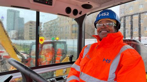 A woman sits in a dumper truck cab. She has hi-viz on and a blue helmet. She is smiling.