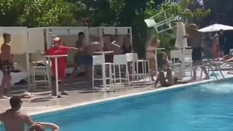 A group stood by a pool as a white chair flies through the air towards a man's head