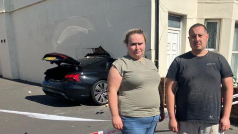 A woman and man stand side-by-side outside of a house with a car buried in the wall