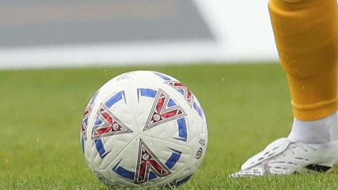 A National League football about to be kicked by a white boot with a yellow sock