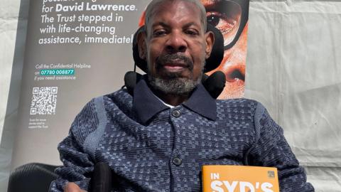 Syd sitting in a wheelchair with a dark polo shirt on, short beard and hair, smiling at the camera. There is a poster behind him and he is outside by a white tent.
