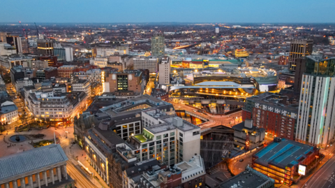 A view of Birmingham city centre taken from the sky. It is early evening as the buildings are visible but the lights and street lights are on. The Rotunda can be seen in the distance in the middle of the shot.