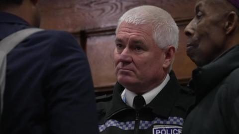A man in a police uniform is talking to members of a church congregation. He has short white hair and is staring directly into the eyes of a man he is talking to.