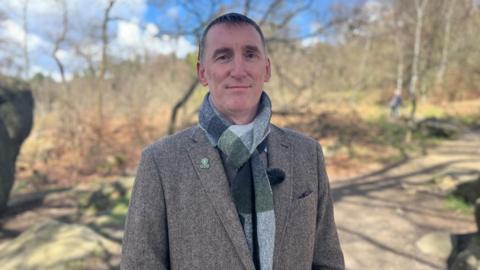 Man in grey tweed jacket stood infront of forest landscape 