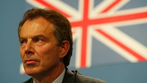 Close-up of Tony Blair at the G8 Summit, looking slightly to the left with a serious expression. He is wearing formal attire, and behind him is a large, blurred Union Jack flag on a blue background.