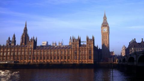 The Houses of Parliament line the river Thames to the left with the Big Ben structure on the right, below a blue sky on a bright day. The glow of the sun is making the gothic brick buildings appear a dark orange.